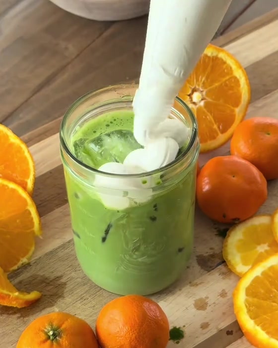Cream being piped onto an iced green matcha with fresh orange slices